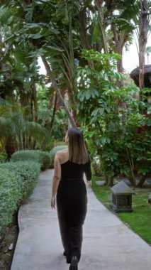 Siyah elbiseli kadın lüks otellerdeki tropikal bahçe yolunda yürüyor. Yeşillik ve palmiye ağaçları sakin bir tatil ortamını vurguluyor..