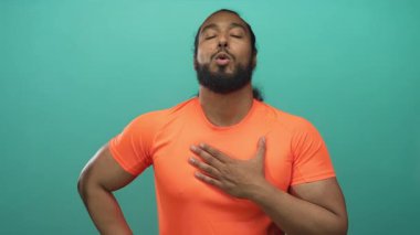 Kalp krizi sırasında göğsüne sarılan turuncu gömlekli bir adam kalp krizi sırasında deniz mavisi arkaplanına dayanır..