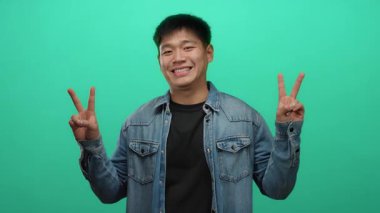 Genç adam rahat kot gömleğiyle gülümsüyor. İki eli de parlak yeşil duvar arkaplanının üzerinde barış işareti yapıyor.