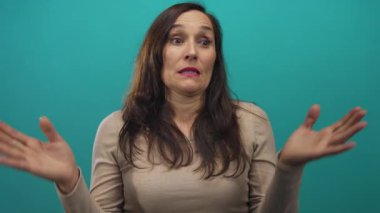 Uzun saçlı bir kadın yeşil arka plan duvarının üzerinden omuz silkip, bej bir kazak giyiyor ve kafa karışıklığını dile getiriyor..