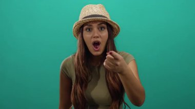 Şapka takan İspanyol kadın şaşkın bir ifadeyle izole edilmiş yeşil arka planı işaret ederek canlı bir zemin üzerinde şaşırtıcı ve meraklı bir şekilde ifade ediyor..