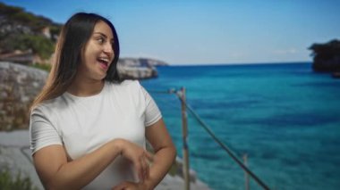 Kadın gülümser ve beyaz gömlek giyerken turkuaz okyanus kıyısındaki korkuluklara parmak basar; keyif daveti.