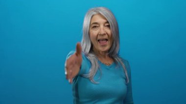 Kadın elini öne uzatıyor, gri saçlı ve gülümsüyor parlak mavi bir arka plana karşı, pozitifliği ve kapalı bir alanda teması iletiyor..