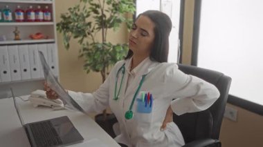 Steteskoplu İspanyol kadın doktor hastane kliniğinde masa ve dizüstü bilgisayarla röntgen çekerken sırt ağrısı çekiyor..
