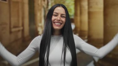 Uzun siyah saçlı, açık havada duran İspanyol kadın, mutlu bir şekilde poz veriyor, beyaz elbiseler giyiyor, sıcak tonlu mimari bir zemine gülümsüyor..