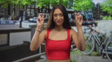Park halindeki bisikletlerin ve kanal korkuluklarının yanında iki eliyle barış işareti gösteren bir kadın, kırmızı bluz ve saat takıyor; iyimserlik..