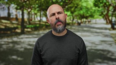 Güneşli bir günde, parkın dışında duran sakallı bir adam yeşil ağaçlarla çevrili, derin düşüncelere dalmış, elleri yüzü kısmen kapalı ve günlük koyu giysiler giymiş bir adam.