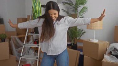 Oturma odasında kutuları, bitkileri ve merdivenleri olan yeni bir evde yerleşmeyi seven bir kadın, günlük kıyafetlerle mutluluğu ifade ediyor, yeni bir başlangıç, neşeli bir yer değiştirme ve rahat bir iç mekan öneriyor..