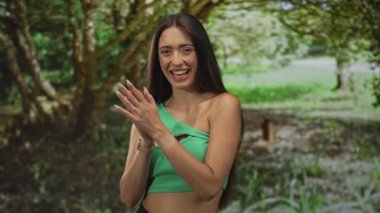 Yeşil ağaçlar ve çimenlerin arasında el çırpan kadın, gülümseyen ve kapanan gözler, yeşil bir omuzda görünen çıplak omuzlar; sevinç kutlamaları.