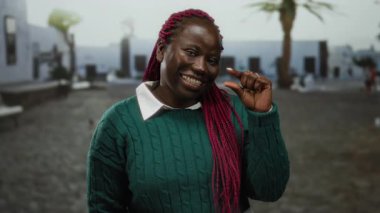 Açık bir şehir caddesinde kırmızı örgülü bir kadın, küçük bir jest yapıyor, yeşil bir kazak giyiyor, arka planda parlak bir şehir ortamını işaret eden palmiye ağaçları var..