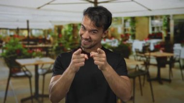 Lokantadaki terasta gülümseyen genç bir adam, dışarıda kahveyle, kameraya şakacı bir şekilde bakıyor, samimi ve davetkar bir ortam yayıyor..