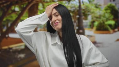 Güneşli bir parkta ağaçlar ve yeşilliklerle gülümseyen bir kadın, beyaz bir gömlek giymiş, rahat ve mutlu, huzurlu bir açık hava anını yakalıyor..