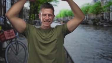 Gündüz vakti Amsterdam caddesinde, gözlüklü ve yeşil tişörtlü bir adam gülümseyip kollarını kaldırıyor..