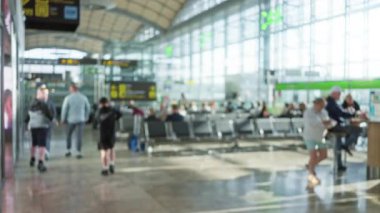 Bulanık insanlar, parlak bir havaalanı terminalinde oturur ve yürür. Güneş büyük pencerelerden süzülür. Parlak modern iç mekan ve tabelalara bokeh etkisi yaratır..