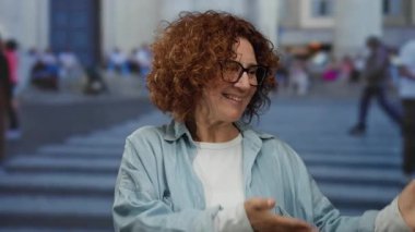 Kalabalık bir caddede el kol hareketi yapan bir kadın, arka planda insanların olduğu bir şehir sahnesini çekiyor..