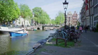 Güneşli bir günde insanların yürüdüğü bulanık bir kanal manzarası, bisikletlerin park ettiği binalar ve şehrin canlı atmosferini yansıtıyor..