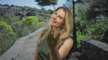 Gülümseyen ve el kol hareketi yapan bir kadın yeşil bir park ortamında, sıcaklığını ve dostluğunu ifade ederek buraya gelir..