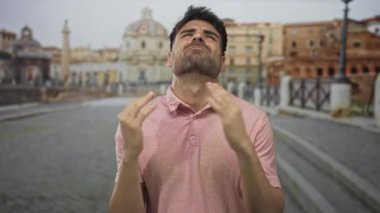 Young hispanic man raises hands on vatican street while looking upward with closed eyes; frustration.