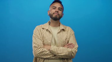 Mavi stüdyoda kolları çaprazlanmış bej rengi gömlekli ve direk bakışlı İspanyol genç bir adam..