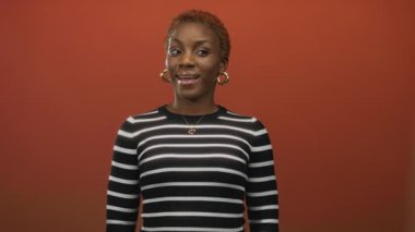 Ağzı açık gülümseyen, halka küpe takan ve çizgili bluzu olan bir kadın stüdyoda turuncu bir duvarın önünde duruyor..