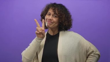 Kadın, geniş bir gülümseme ile canlı mor bir stüdyo arkaplan karşısında eli ile iki parmak barış işareti sunar; sevinç.