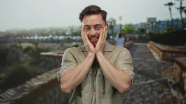 Sokak ortasında gülümseyen sakallı genç bir adam şehir arkaplanına karşı elleriyle yüzlerine dokunuyor.