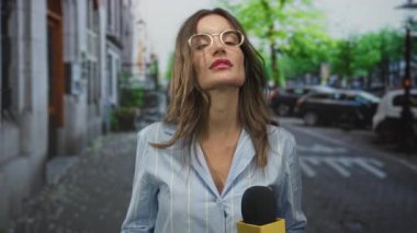 Sokakta konuşurken elinde mikrofon tutan kadın muhabir avuç içi kaldırma hareketleri; acil haber bildirimi.