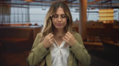 Gözlük takan iş kadını, restoranda saç ve pürüzsüz ceket yakasını düzeltiyor; kendine güven güçlendiriyor.
