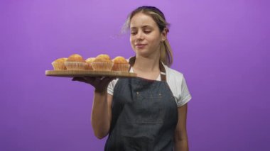 Mor bir stüdyoda ahşap bir tepsi kek tutarken gülümseyen genç kadın fırıncı; ev yapımı pişirme tatmini.