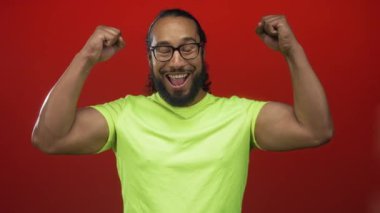 Kırmızı bir stüdyoda kaslarını esneten ve gülümseyen bir adam hem de yumrukları havada, gözlüklü neon yeşil bir gömlek giyerken; neşe zaferi..