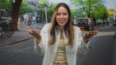 Beyaz tişörtlü ve sarı kolsuz tişörtlü bir kadın işlek bir caddede ağaçlar ve arabalarla omuz silkip el kaldırıyor..