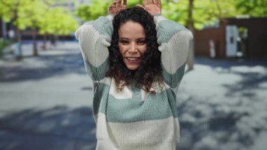 Genç bir kadın, güneşli bir günde arka planda ağaçlarla dışarıda dikilirken elleri kafasında tavşan kulağı hareketi yapıyor..