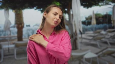 Pembe tişörtlü kadın, dışarıda bir tatil havuzunda, palmiye ağaçları ve dinlenme yerleriyle omuz ağrısı çekiyor. Tatil ortamında huzursuzluk ve sükuneti simgeliyor..