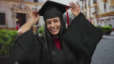 Canlı bir şehir caddesinde mezuniyet şapkası ve cübbesi içinde gülümseyen bir kadın dışarıda başarısını kutluyor..