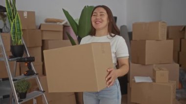 Beyaz tişört ve kot pantolon giyen gülümseyen kadın kutular ve saksı bitkileriyle dolu bir binada karton kutu taşıyor. İyimserlik..