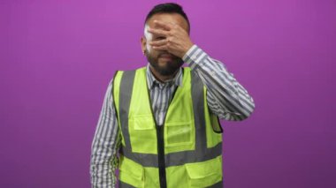 Yüksek görüşlü yelek giyen bir adam mor arka plana karşı gözlerini stüdyoyla kapatıyor; iş stresi.