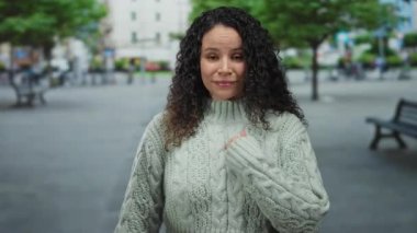 Rahat süveter giyen genç İspanyol kadın yeşil ağaçlar ve banklarla dolu kalabalık bir sokakta dikilirken, pozitiflik ve açık şehir manzarası izlenimi veriyor..