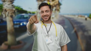 Sakallı ve steteskoplu genç adam arka planda palmiye ağaçları ve okyanusla güneşli bir deniz kenarında kameraya gülümsüyor..