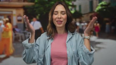 Şehir caddesinde kot ve pembe giyen bir kadın, neşeli bir ifadeyle gözlerini kapatıyor, açık havada insanlarla çevrili, canlı ve canlı şehir manzarası, İspanyol kökenli.