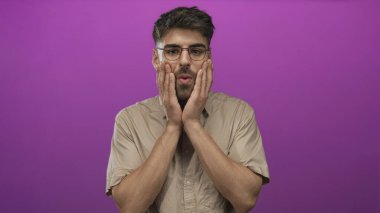 Gözlüklü ve bej gömlekli bir adam mor stüdyoda iki elini açık ağızla yanaklarına bastırır ve kaşlarını kaldırır..