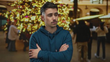 Çapraz kollu genç İspanyol bir adam, canlı bir akşamda düşünceli bir ruh hali somutlaştıran, parlak tatil ışıklarının şenlikli bir arka planına karşı bir şehir caddesinde duruyor..