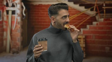 Waffle ısıran ve gri balıkçı yaka kazaklı kahve fincanı tutan bir adam inşaat binasının içinde; rahatlık düşkünlüğü.