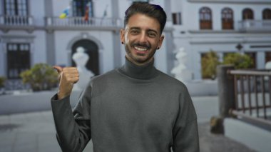 Gülen adam, beyaz bina balkonunun önündeki güneşli şehir caddesini işaret ediyor; dostça bir davet.