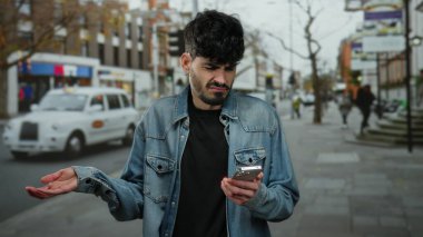 Akıllı telefona bakan genç adam, şehir sokaklarında kafası karışmış bir halde, şehir geçmişi bulanık kot ceket giyiyor, trafik ve binalar var..