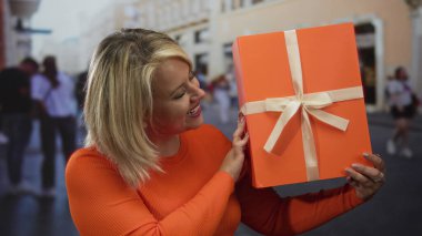 Kalabalık şehir caddesinde elinde büyük turuncu hediye kutusu olan bir kadın..