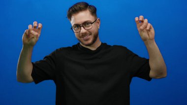 Sakallı ve gözlüklü genç adam, iki eliyle barış işareti yapıyor. Mavi arka planda, sakin ve dost canlısı bir tavır sergiliyor..