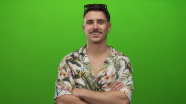 Çapraz kollu İspanyol genç adam çiçekli tişörtle izole edilmiş yeşil arka planda tatilin tadını çıkarıyor, rahat bir turist havası yayıyor..