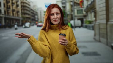 Elinde kahve fincanı tutan kadın kentin caddesinde düşünceli, sarı kazaklı, kızıl saçlı ve bulutlu gökyüzü olan bir kadın..