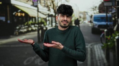 Kentsel caddelerde omuz silken bir adam, şehir ortamında gündelik tarz ve eğlence ifadesiyle ayakta dururken kargaşa ya da belirsizlik taşır.