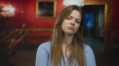 Kollarını kavuşturmuş ve dudaklarını bükmüş bir kadın sanat galerisinde çerçeveli resimler ve avizeyle duruyor..